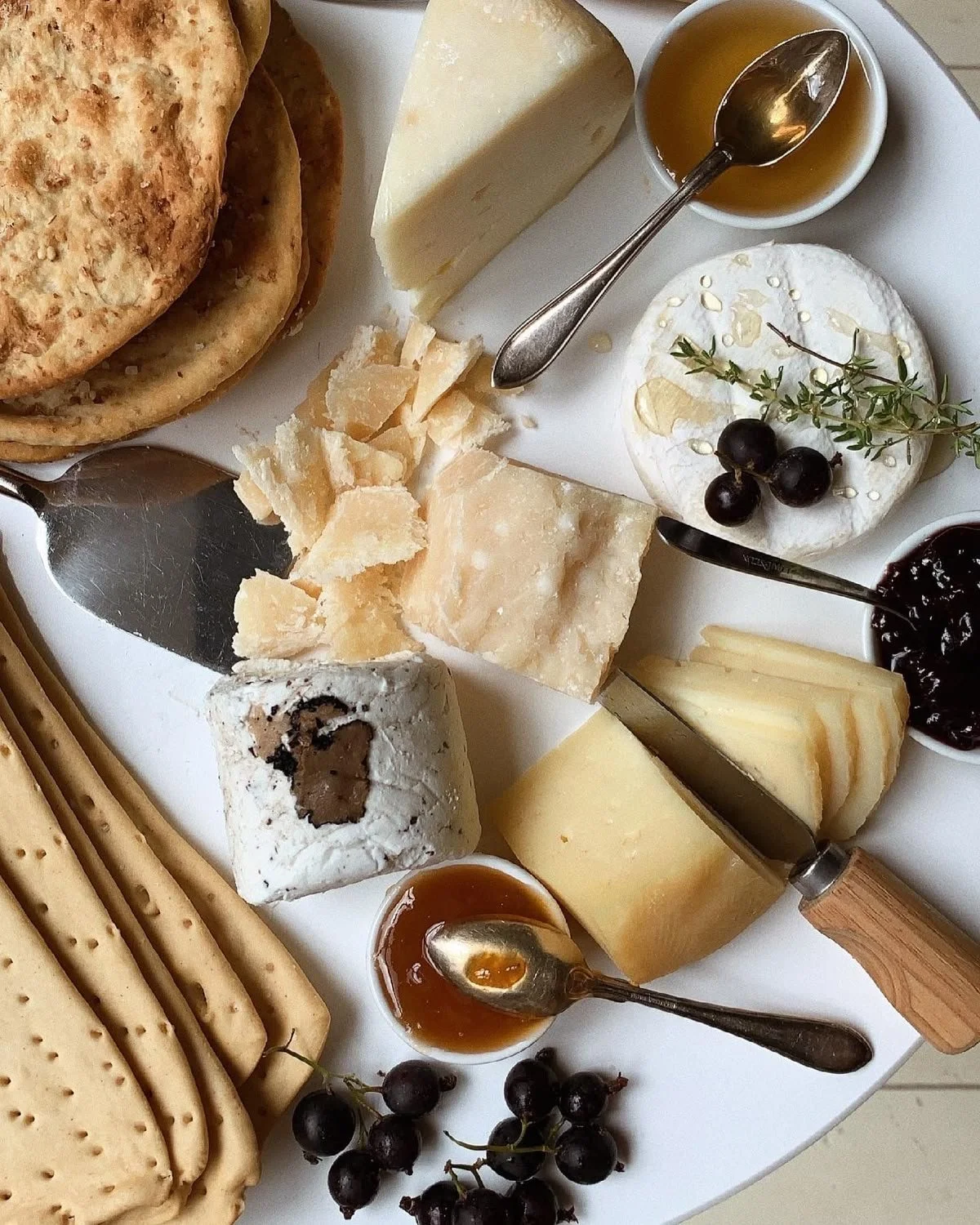 Plate de fromages variés, crackers, confitures, miel, fruits noirs, et pain, avec des cuillères pour les dégustations.