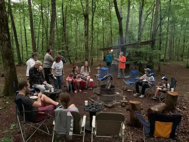 People gathered around a campfire in a wooded area, some sitting on chairs and others standing, with a small shed in the background.