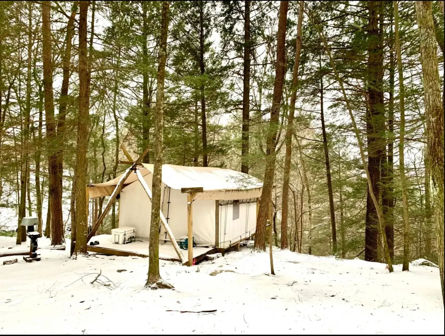 A white winterized tent set up in a snowy forest with tall trees surrounding it.