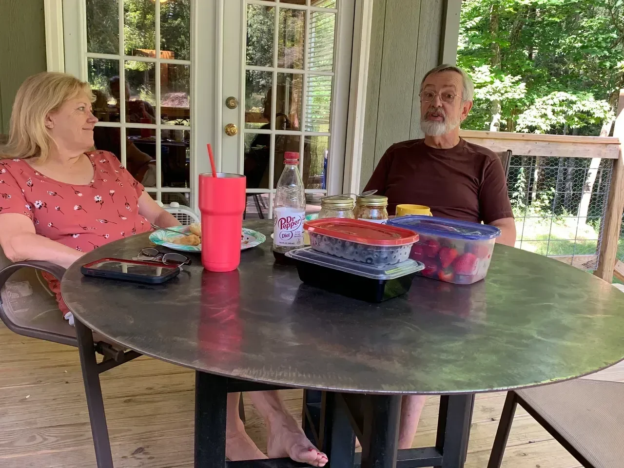 An elderly couple sitting at a round outdoor table with food and drinks, having a conversation on a porch with trees visible outside.
