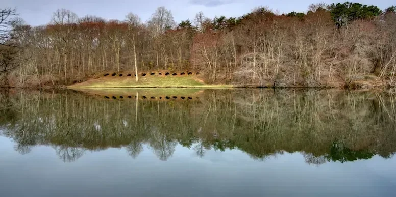 A lake with a wooded shoreline and a grassy area with large drainage pipes or culverts on the hill.
