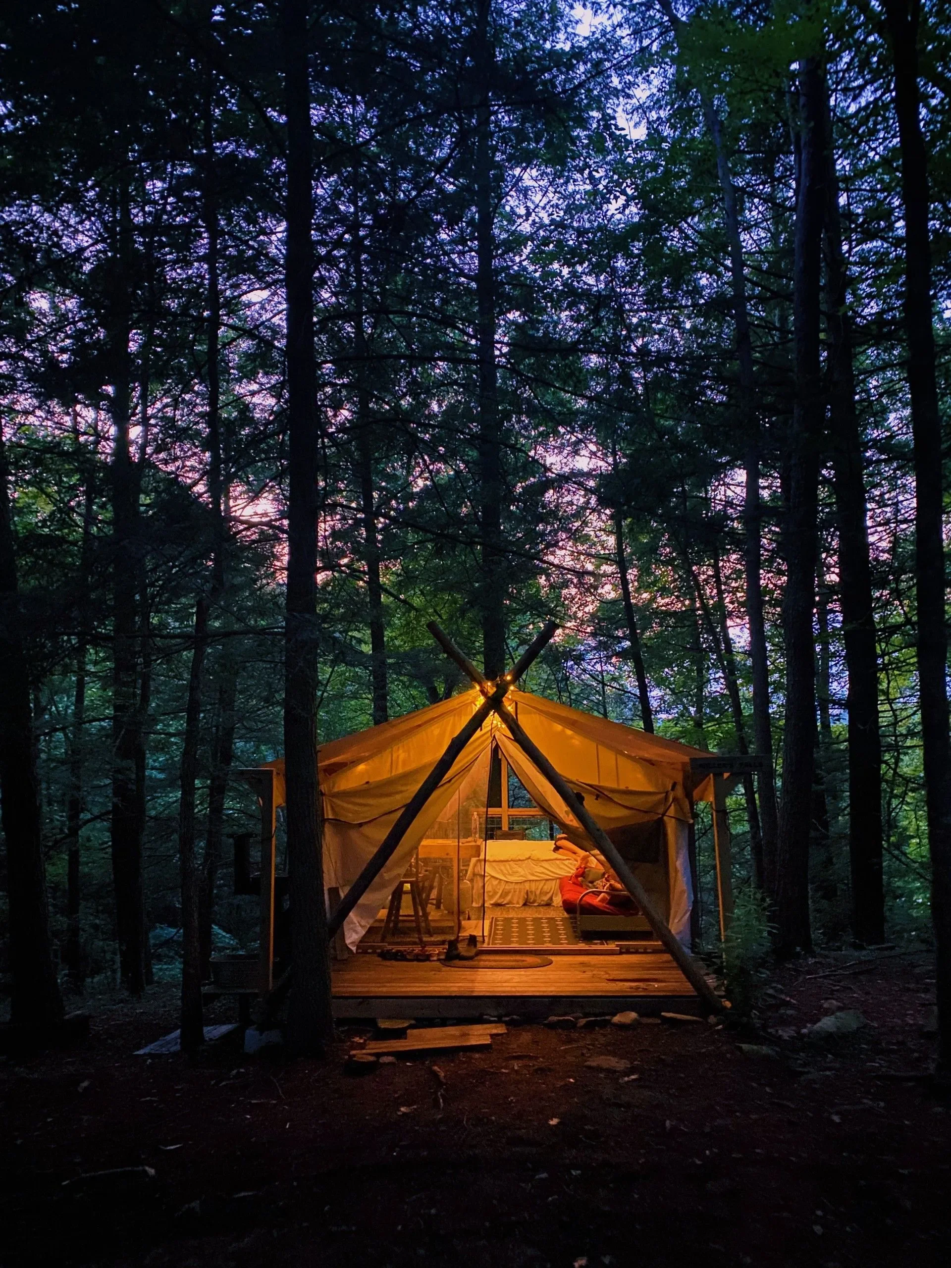 A cozy tent with warm interior lighting set up in a dense forest during dusk, with tall trees and a purple and pink sky in the background.