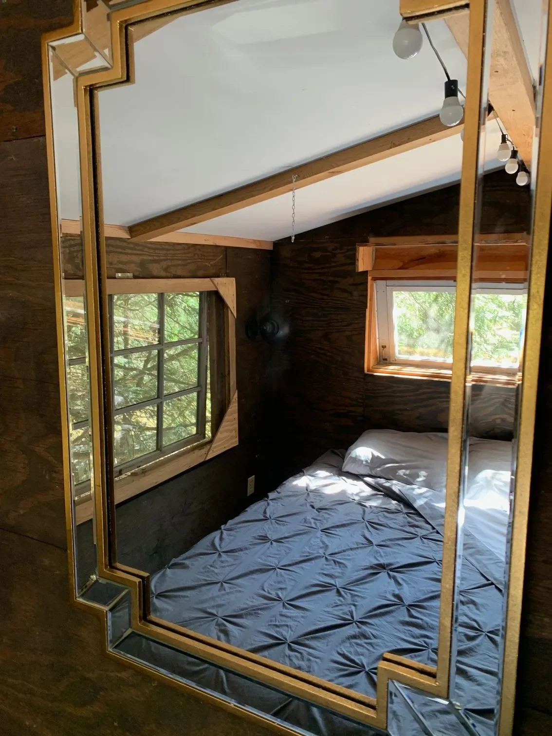 Reflection of a bedroom with wood-paneled walls, a bed with a grey quilt, two windows with wooden trim, and string lights, seen in a mirror with a gold and mirrored border.