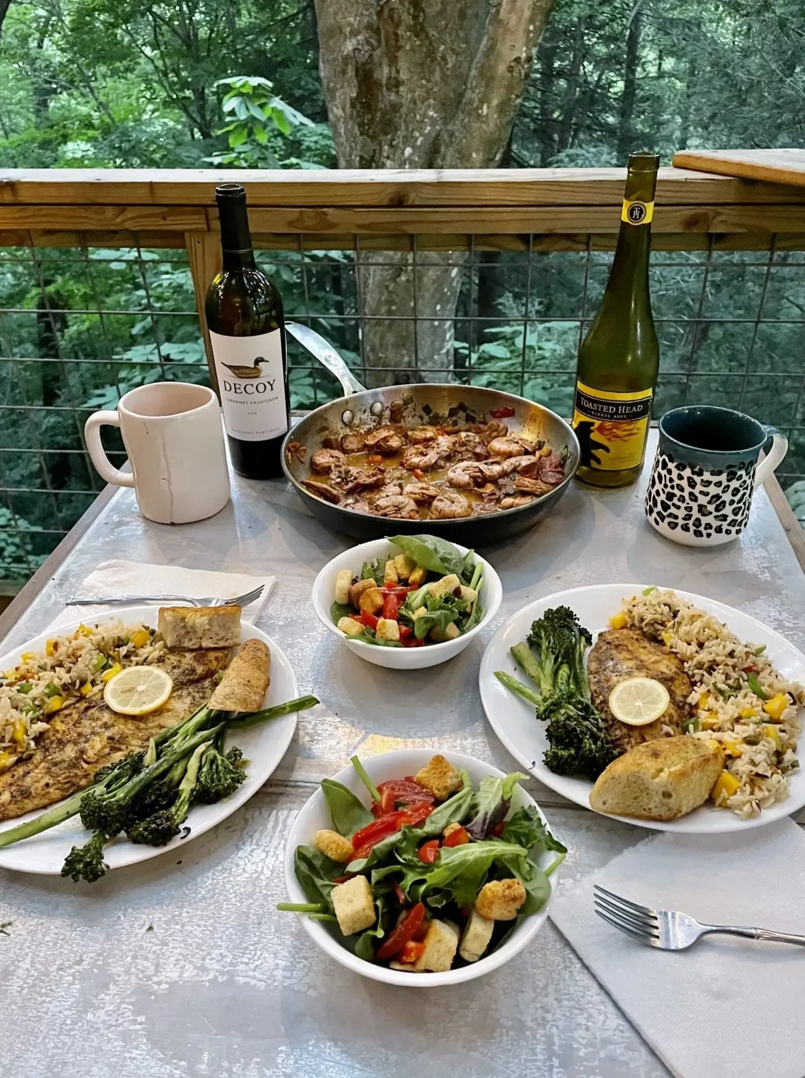 A table set for an outdoor meal with two plates of rice and chicken, a salad, a bowl of fresh greens, a skillet with cooked chicken and vegetables, two bottles of wine, and two coffee mugs against a green wooded background.