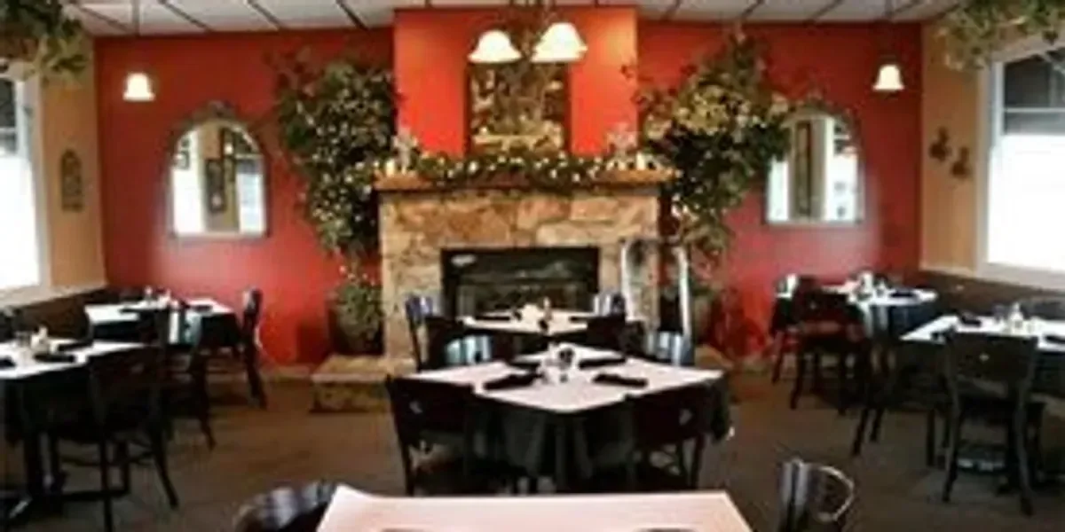 Restaurant interior with red walls, stone fireplace, black tables with white tablecloths, and black chairs, decorated with greenery and string lights.
