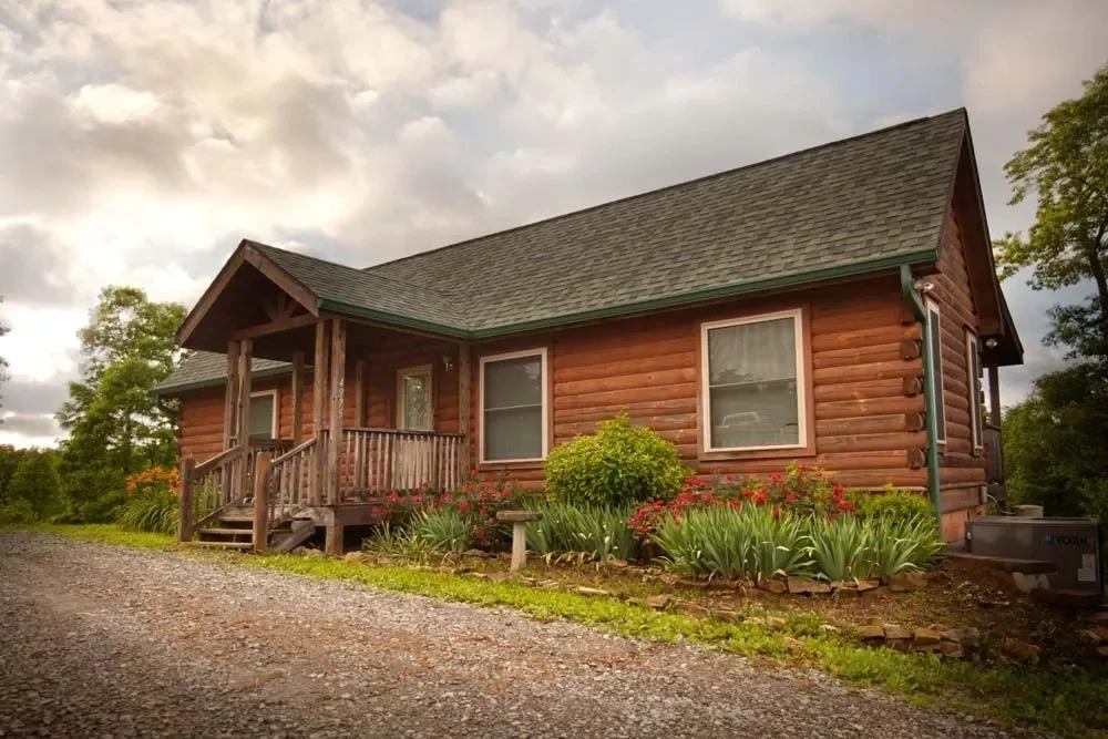 A wooden log cabin with a front porch, surrounded by colorful flowers and greenery under a cloudy sky.