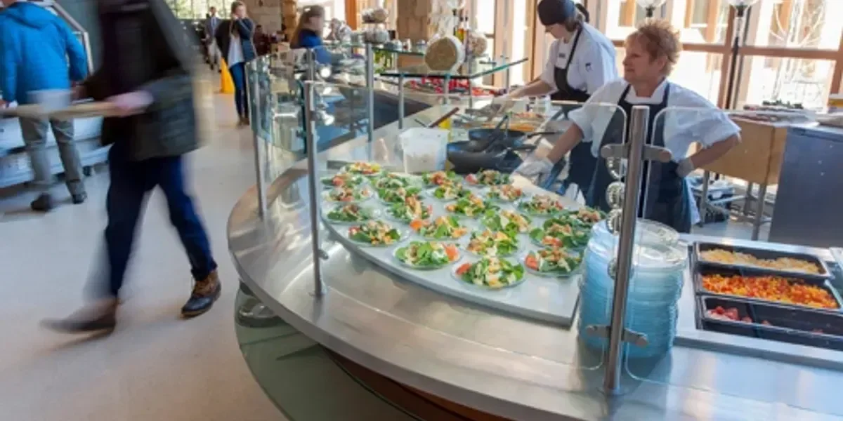 Buffet station with salads and toppings, chefs preparing food, and people serving themselves in a well-lit dining area.