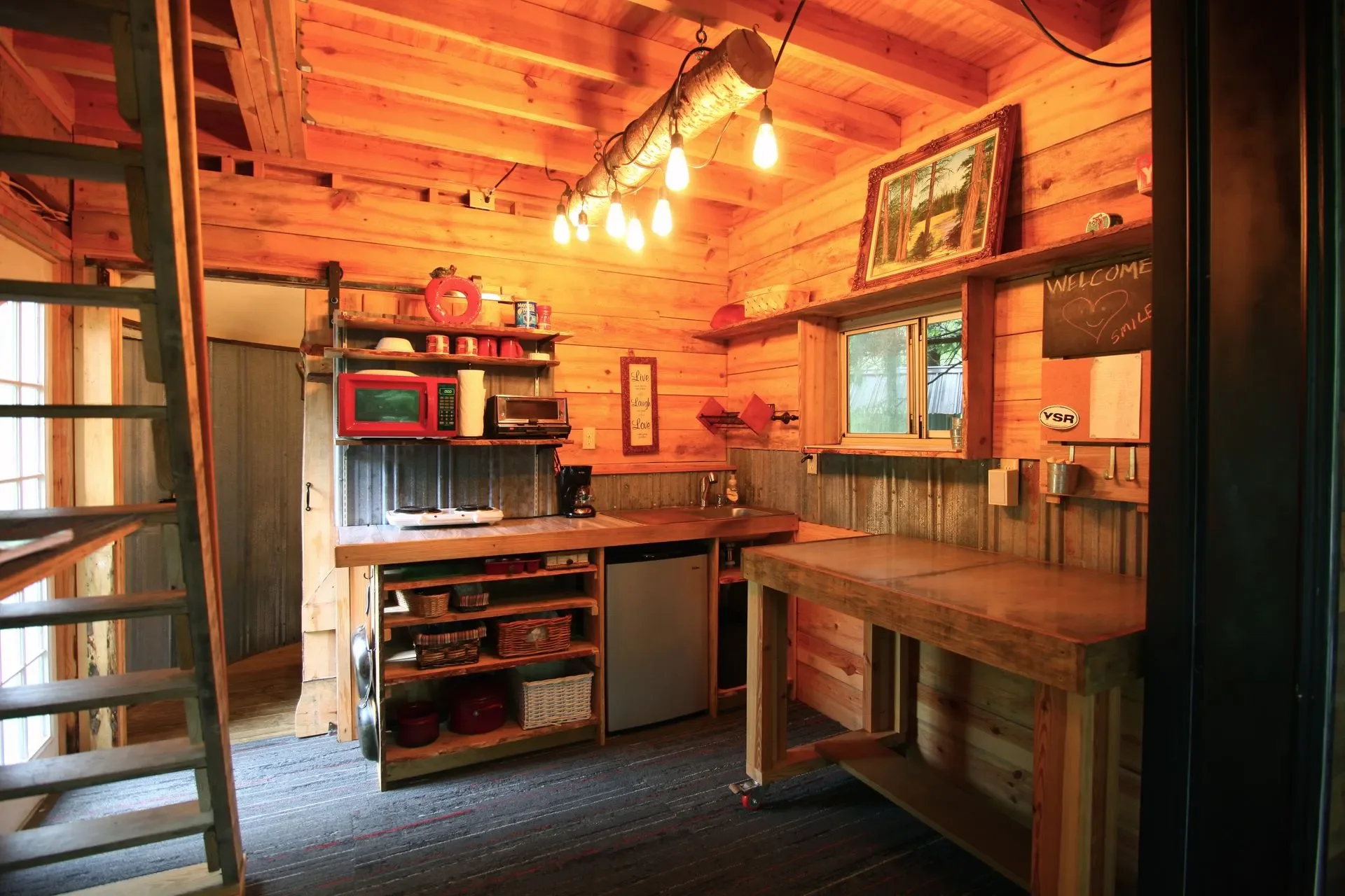 Inside a rustic kitchen with wooden walls and ceiling, featuring a small sink, countertop, microwave, toaster oven, coffee maker, various baskets and containers, a window, framed artwork, a chalkboard with a welcome message, and a hanging light fixtu