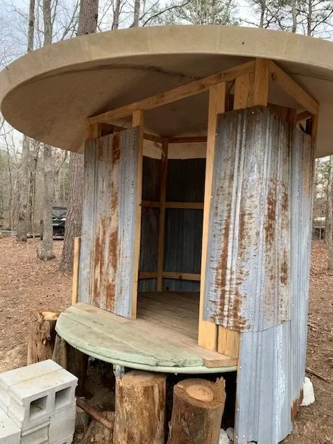 Small outdoor wooden structure resembling a playhouse or treehouse, built on a tree stump with a round concrete roof and corrugated metal walls.