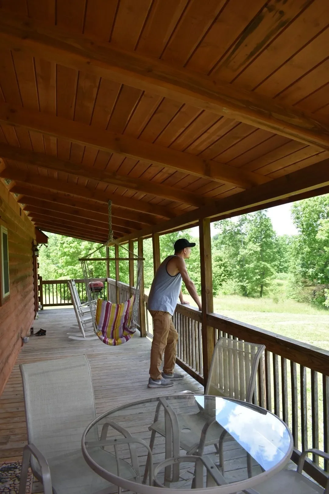 A man wearing a white tank top, khaki pants, gray shoes, and a black cap looks out over a green yard from a wooden porch. The porch features a round glass-top table with four chairs, a hanging swing with colorful striped cushions, and a charcoal gril