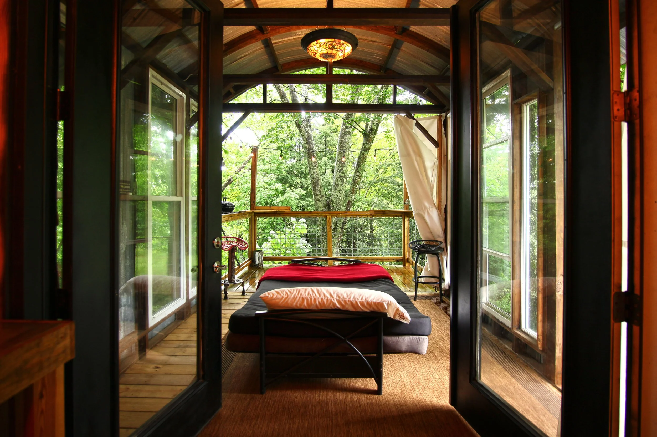 View from inside a cozy rustic bedroom with glass double doors opening to a screened porch surrounded by green trees, with a bed and pillows inside.