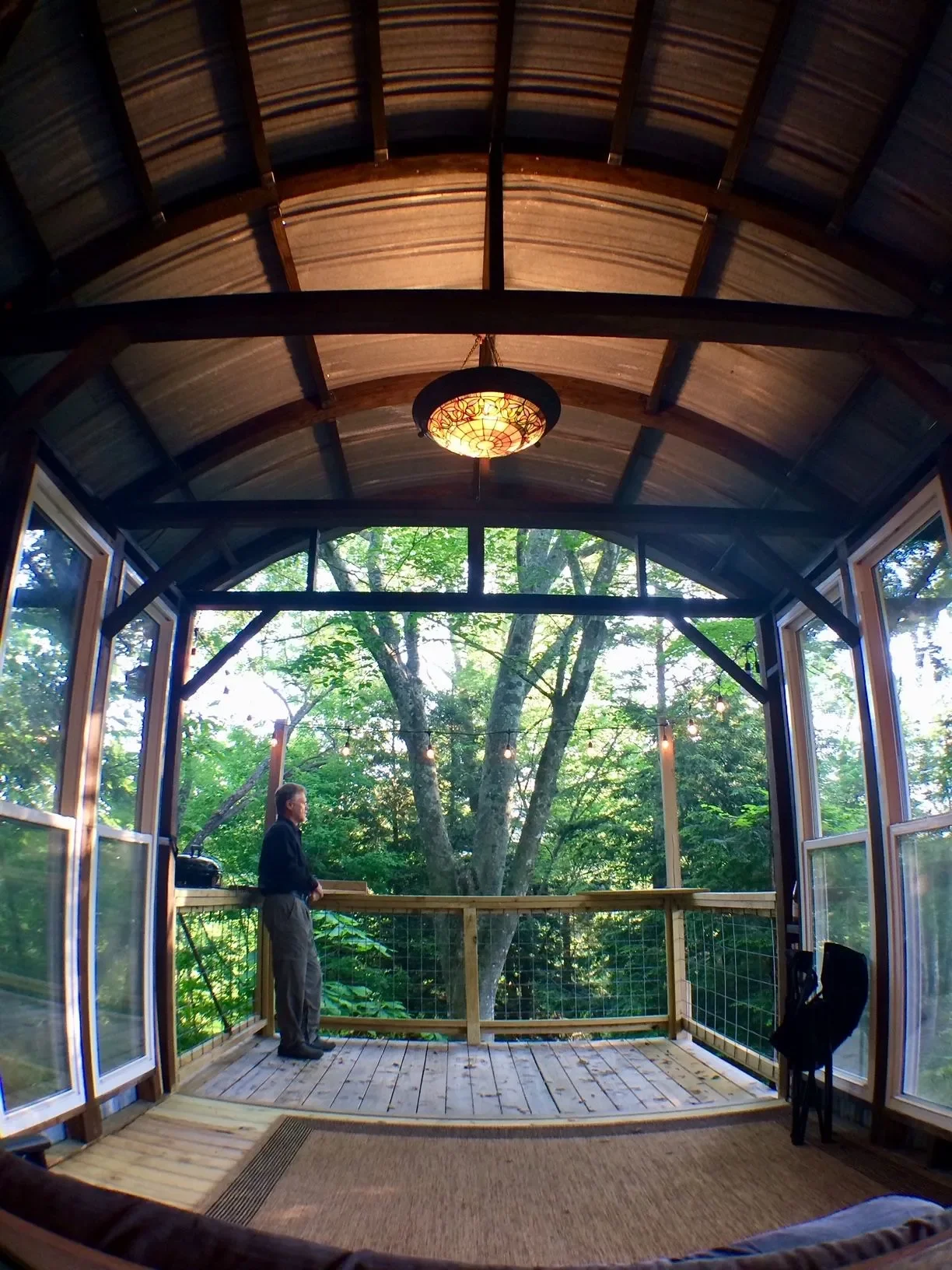 Man standing on wooden deck in a room with large open windows, overlooking a lush green forest with trees, under a vaulted wooden ceiling with a hanging light fixture.