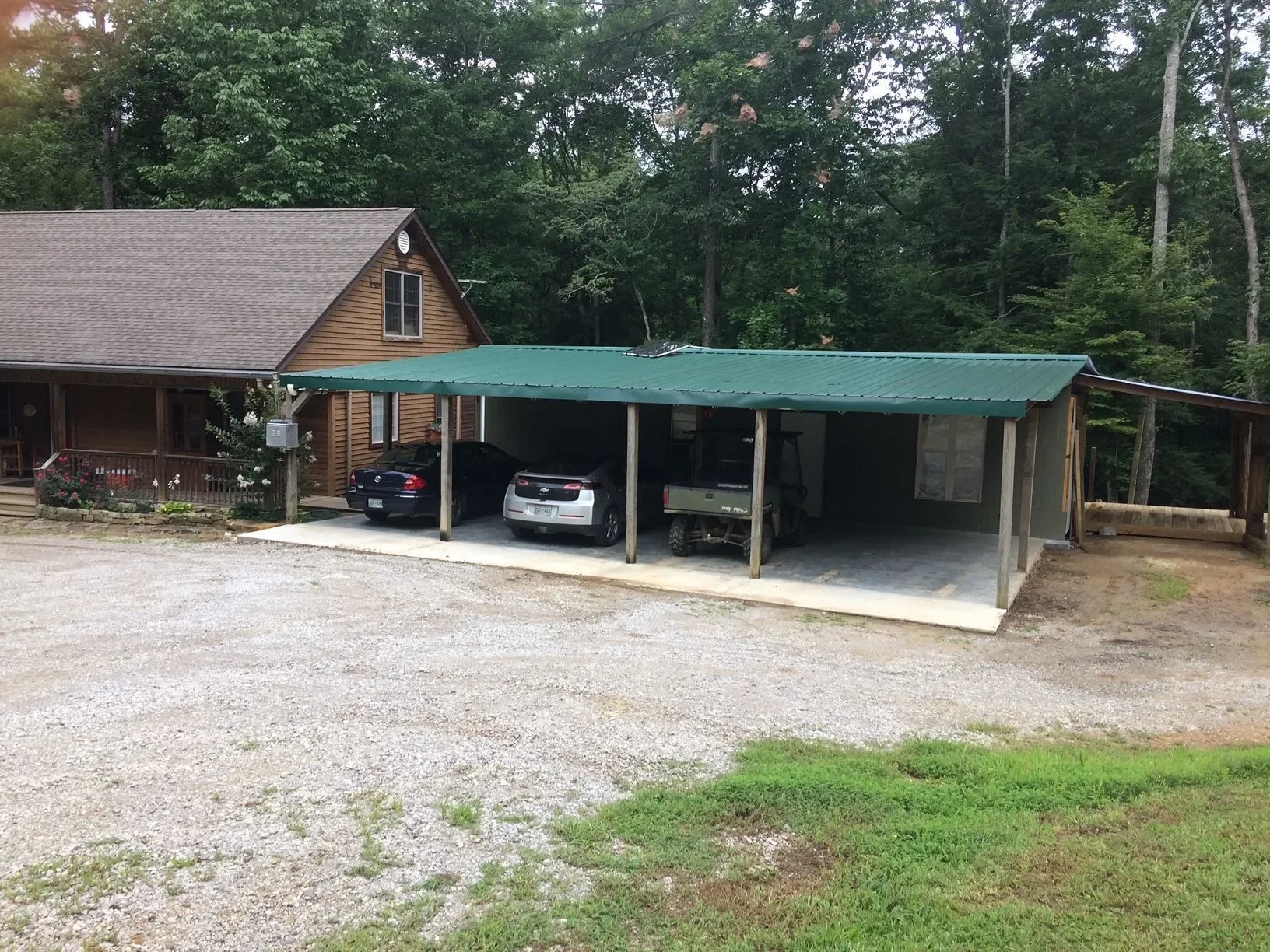 A house with a porch and a small garden, next to a carport with three vehicles parked under a green metal roof, in a wooded area.