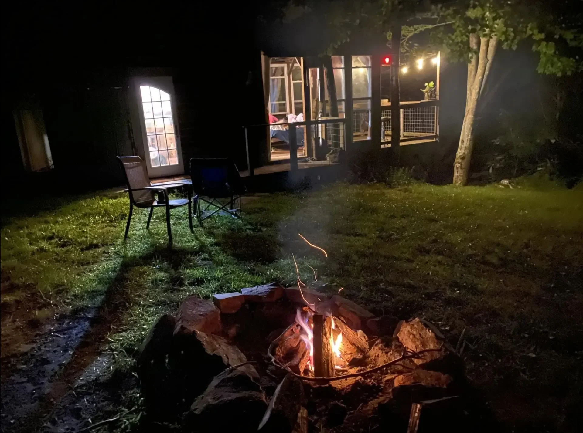 Nighttime outdoor scene featuring a small campfire with glowing embers and flames on the grass. In the background, there is a dark house or cabin with lit interior lights visible through large windows and a door, along with outdoor chairs near the fi