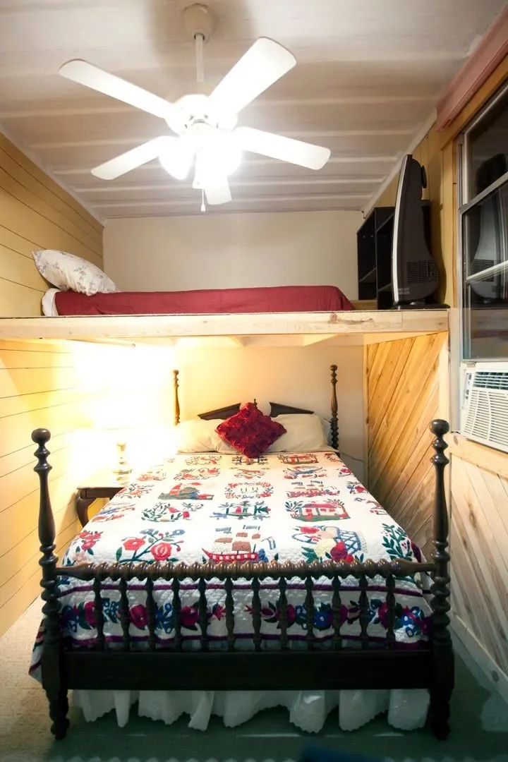 Room with a double bed in front of the camera, topped with Christmas-themed quilt and a red decorative pillow. To the left, a bedside table with a lamp. Above the bed, a bunk bed with a red mattress and pillow. The ceiling has a white fan with five b