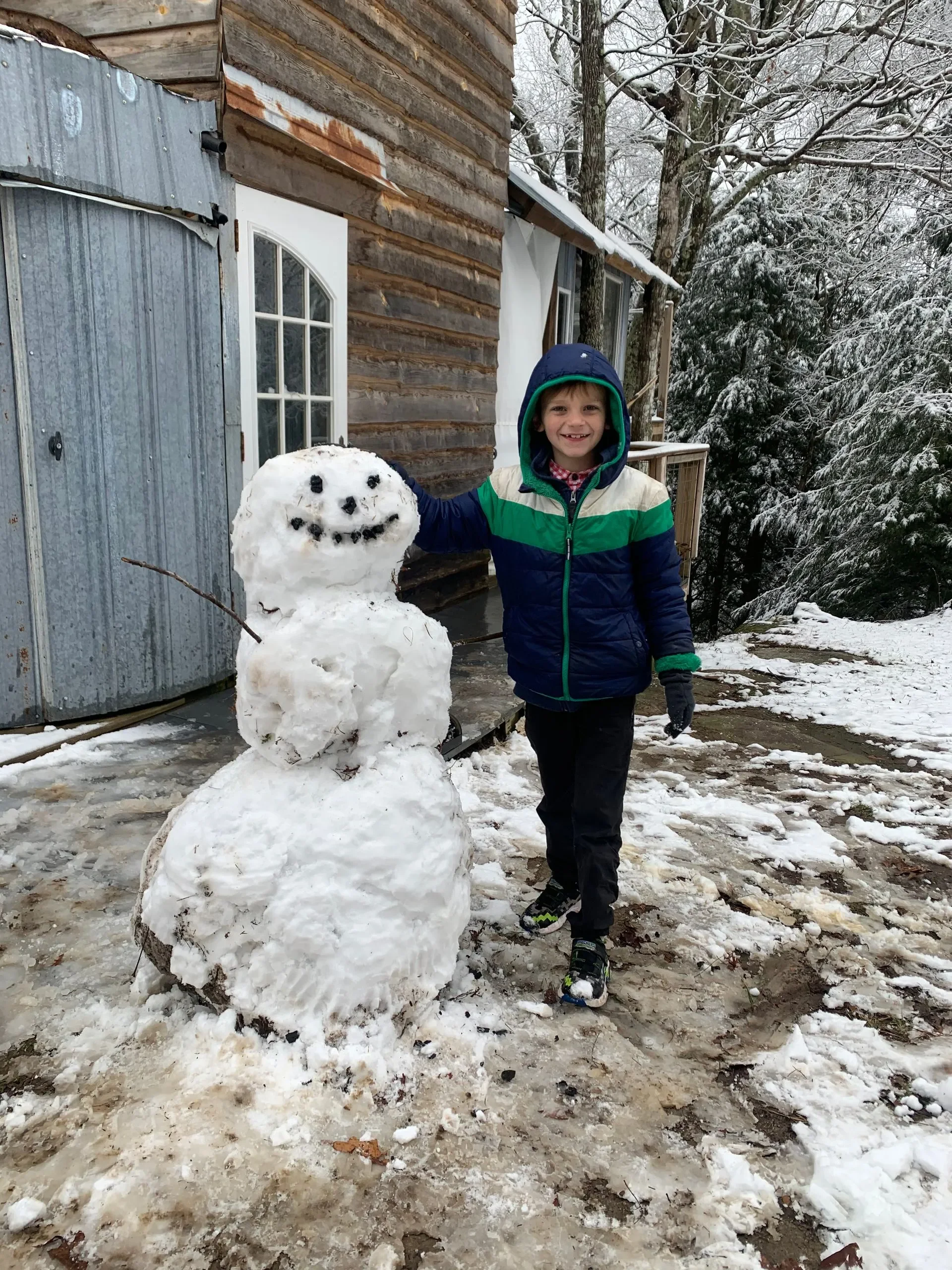 A young boy in a blue, green, and white jacket stands outside on snow-covered ground, smiling and pointing to a small snowman with a smiling face made of coal or similar black material and a stick arm, next to a rustic wooden building and snow-covere