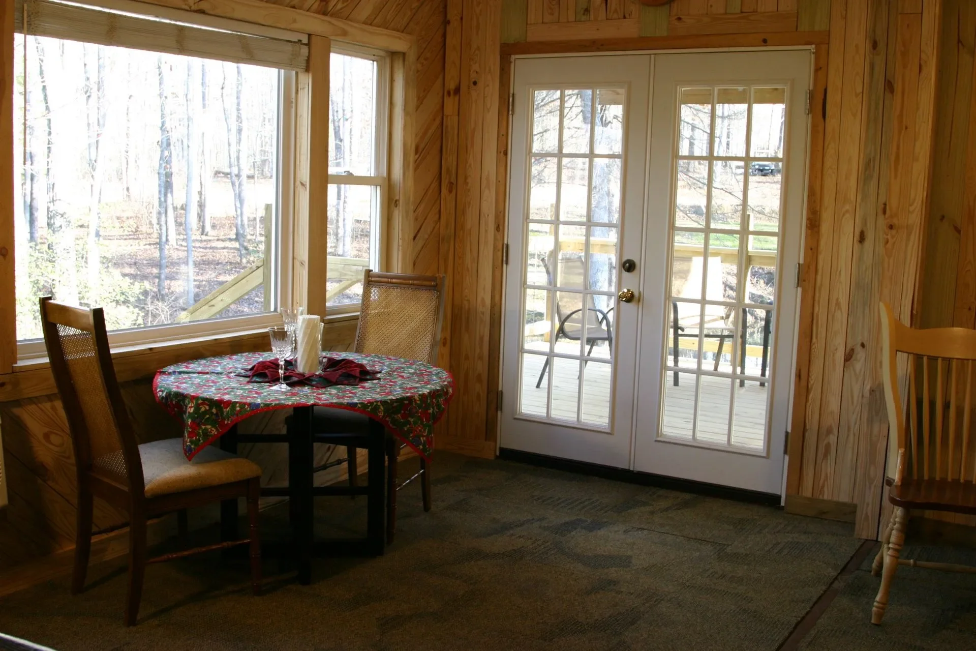 A cozy dining area with a round table covered in a Christmas-themed tablecloth, set with wine glasses and napkins, next to large windows and a glass door leading to an outdoor deck with patio chairs, surrounded by trees.