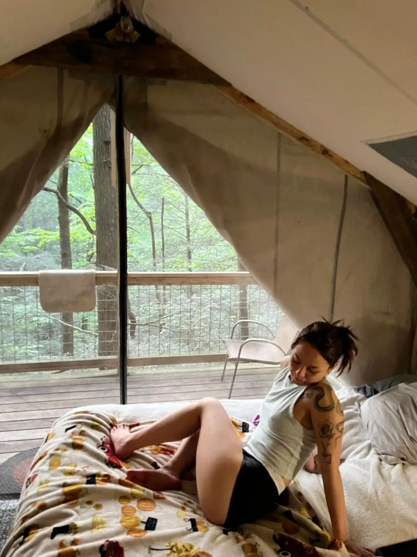 A woman with tattoos on her arm sits on a bed inside a cozy cabin with a sloped ceiling. She is looking down and has her hair tied up. The cabin has a large window showing a view of trees and a balcony with a chair outside.