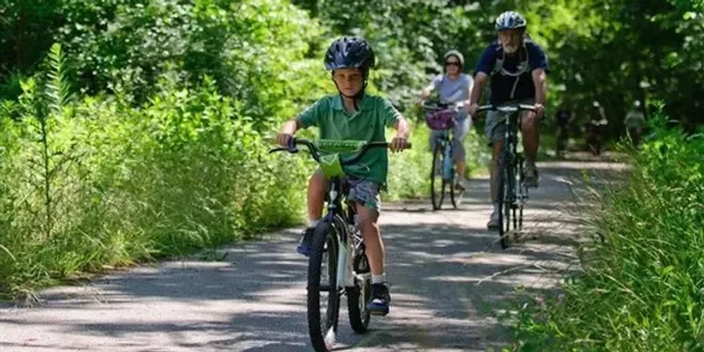 Children and adults riding bicycles on a tree-lined path in a park.