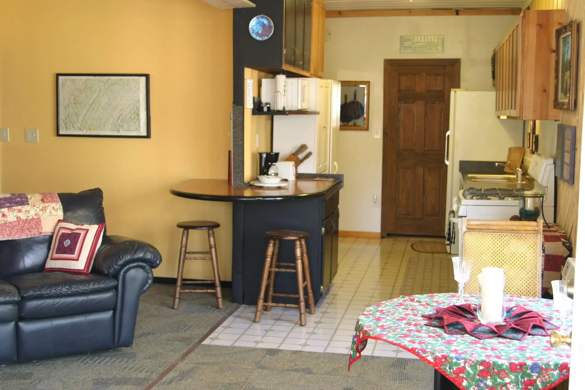 A cozy kitchen and living area with a black leather sofa on the left, a circular dining table with a Christmas-themed tablecloth and candles on the right, a small kitchen with a white stove, cabinets, a sink, and a wooden door at the back. Bar stools