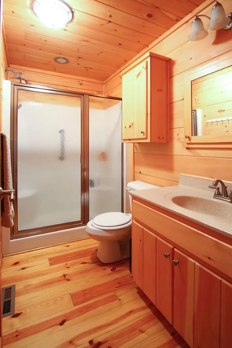 A small bathroom with wooden walls, ceiling, and floor, featuring a glass shower enclosure, a toilet, a wooden vanity with a mirror, and a cabinet above the toilet.