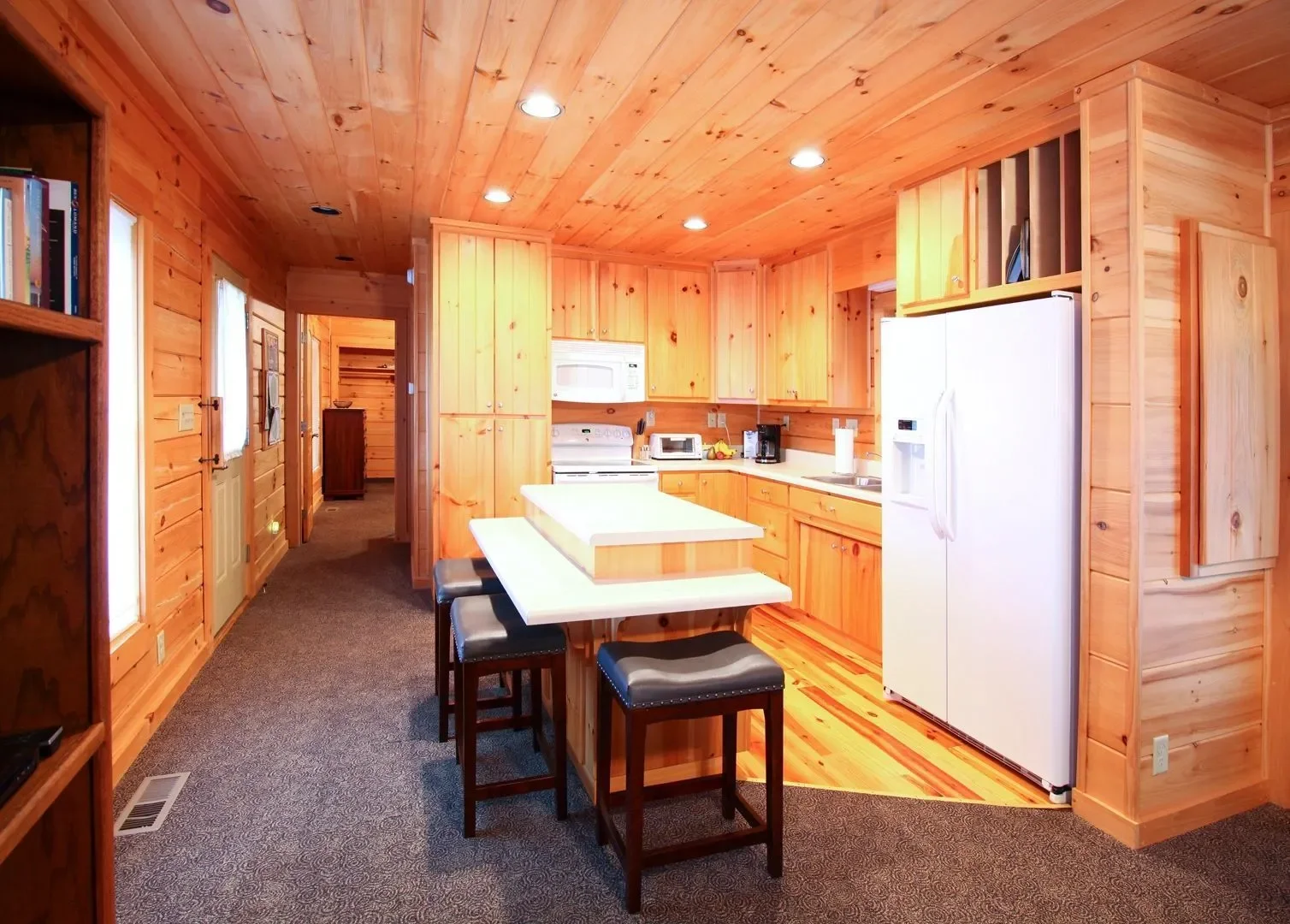 Wooden kitchen with white appliances, black stools, and a small island, in a cabin-style home.