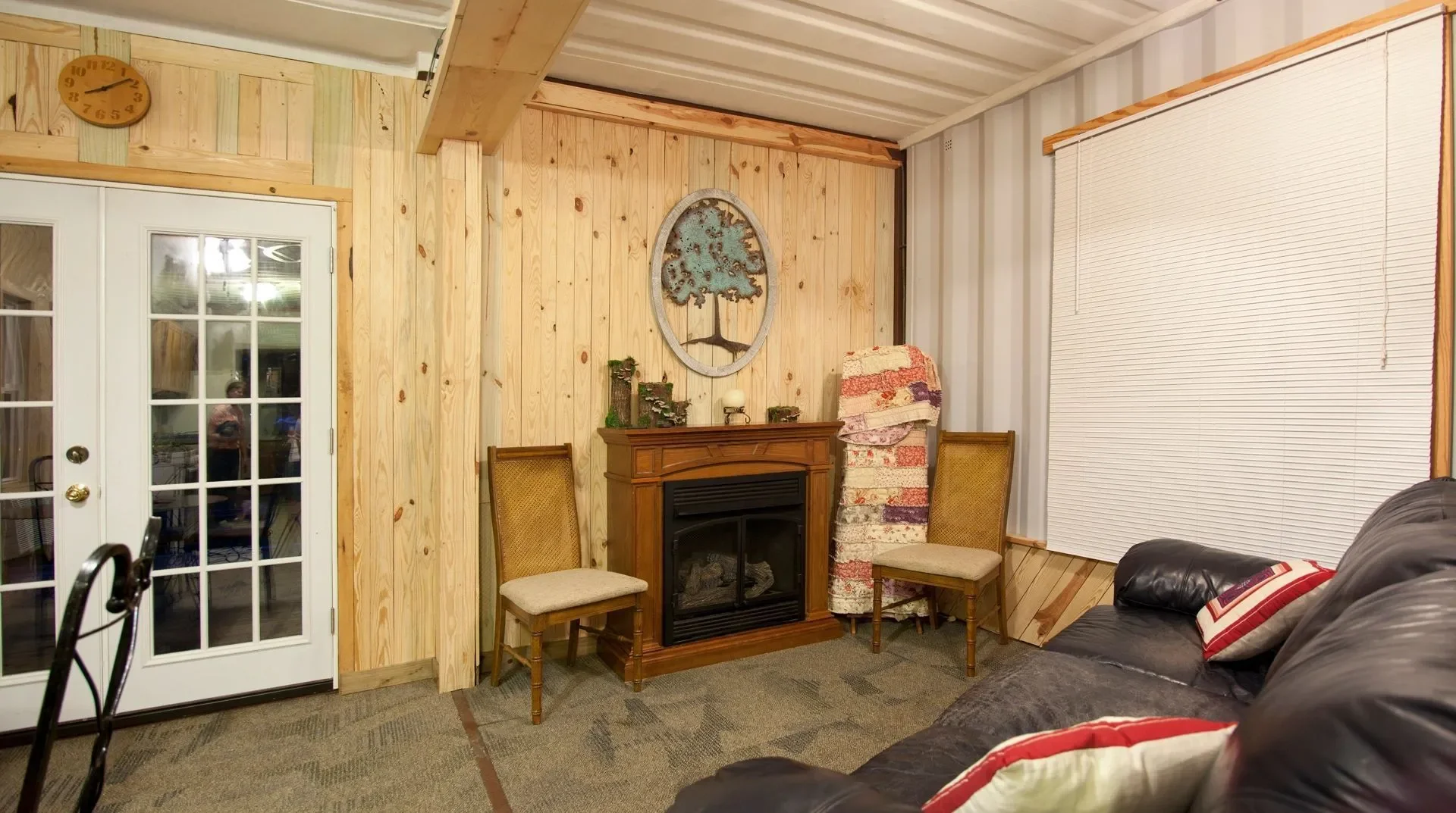 Living room with wood-paneled wall, a fireplace, two chairs, a rolled-up rug, a window with closed blinds, and a dark leather couch with pillows.