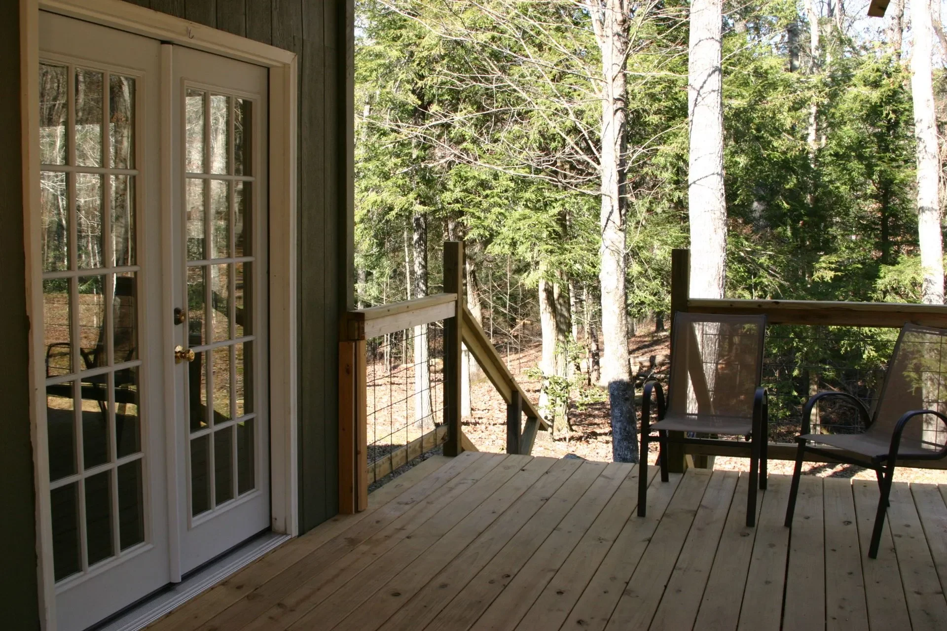 View of a wooden deck with two chairs, overlooking a wooded forest area with tall trees and green foliage.