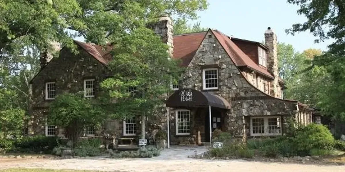 A large stone house with a red roof and multiple chimneys, surrounded by green trees and shrubbery, with a bench near the entrance.