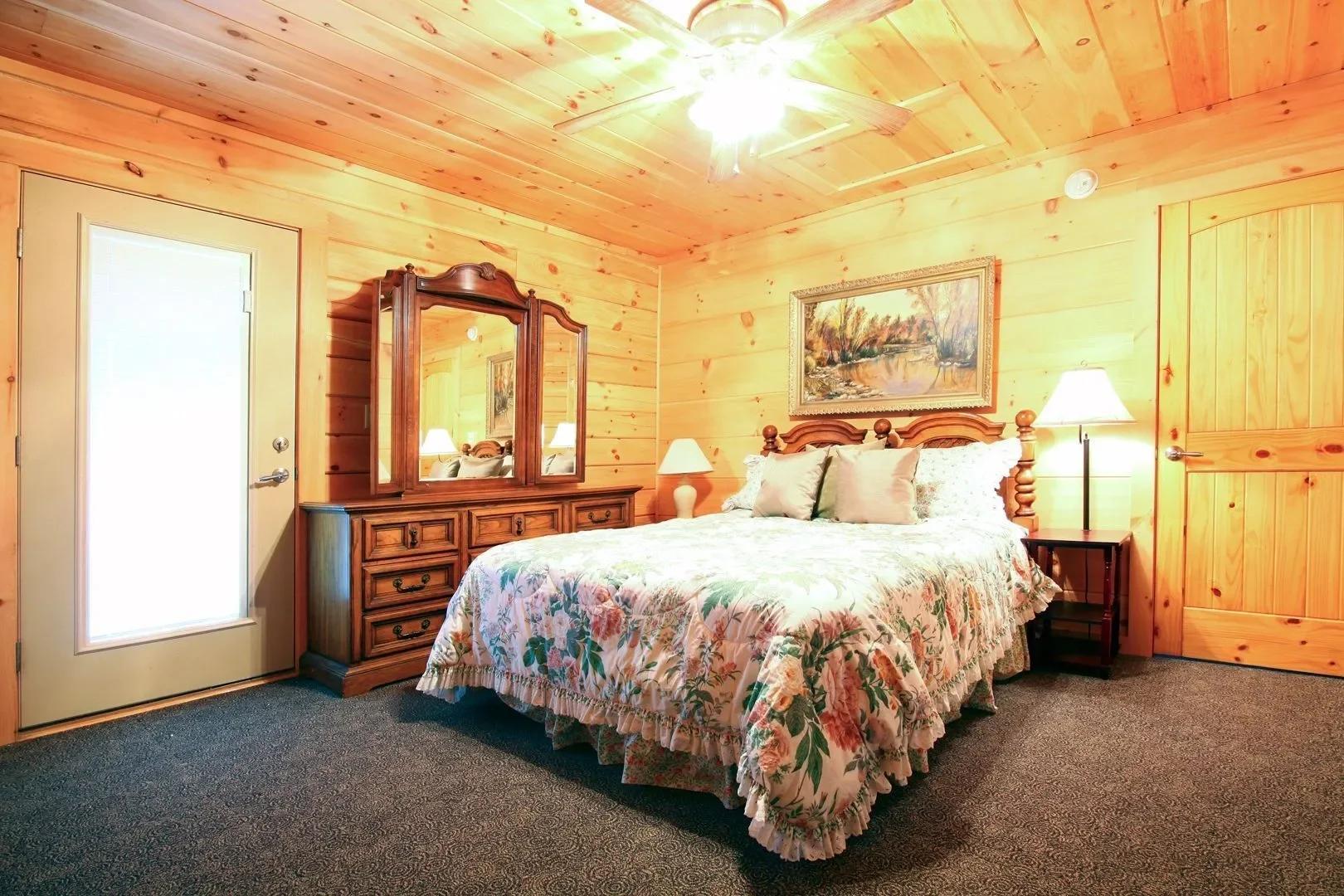 A cozy bedroom with wood-paneled walls and ceiling, featuring a bed with floral bedding and multiple pillows. There is a wooden dresser with a mirror on top, a nightstand with a lamp, and a framed landscape painting above the bed. The room has carpet