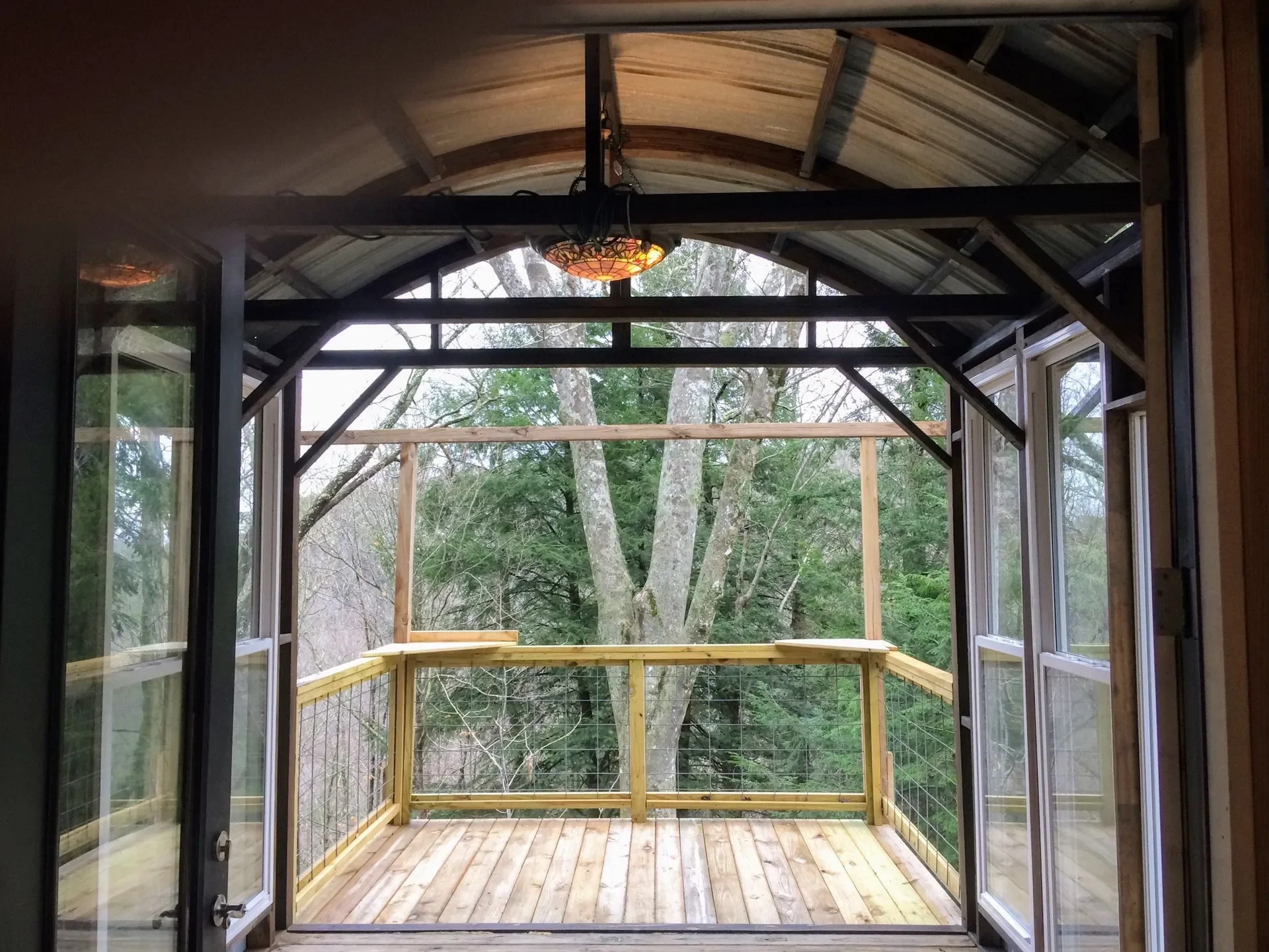 View of a newly built wooden deck with safety railing, outdoor trees in the background, and a ceiling light fixture in the enclosed porch.