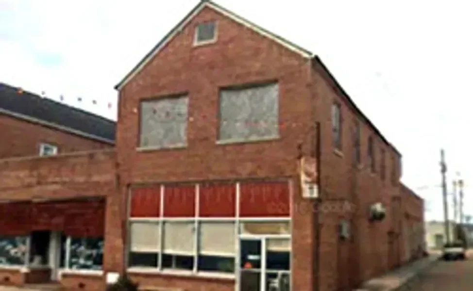 A two-story brick building with boarded-up windows on both floors and a storefront on the ground level with broken or covered windows.