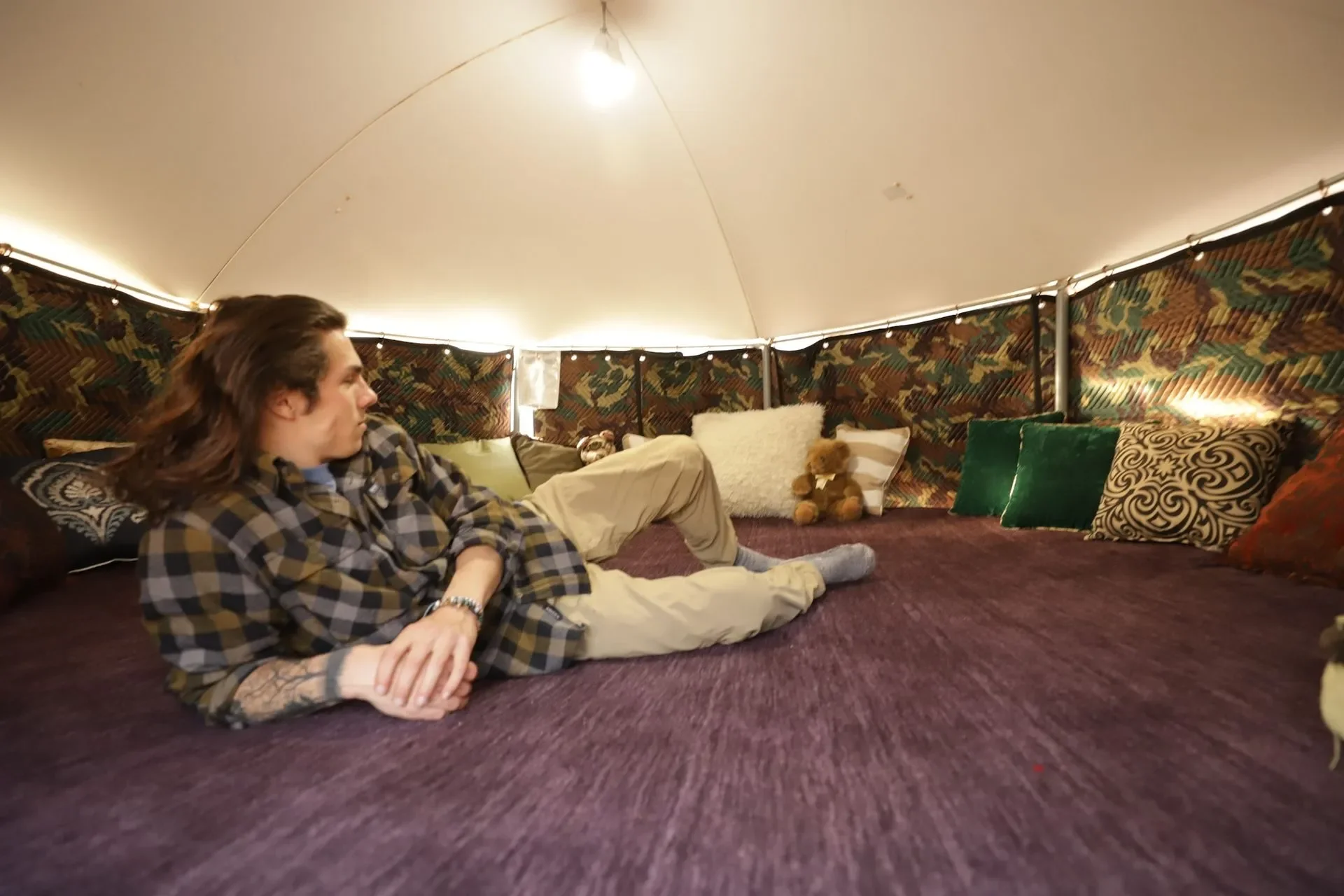 Young man with long hair lying on his side inside a cozy, decorated tent with colorful cushions and plush toys.