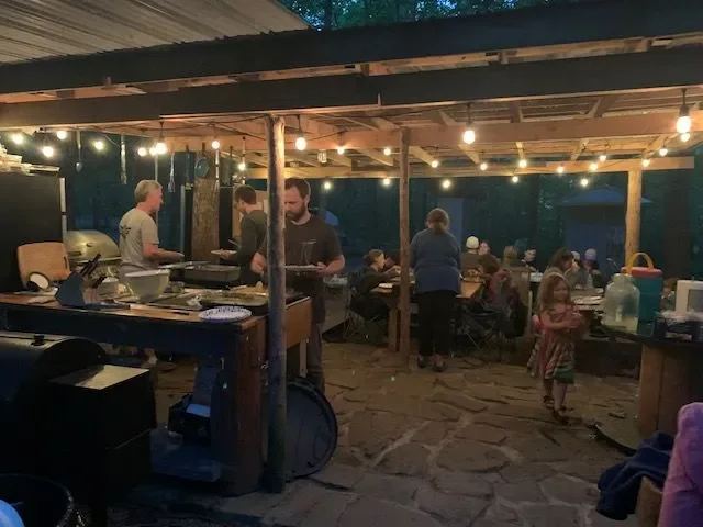 People gathered at a casual outdoor dining area with string lights, wooden beams, and a stone floor, eating and socializing.