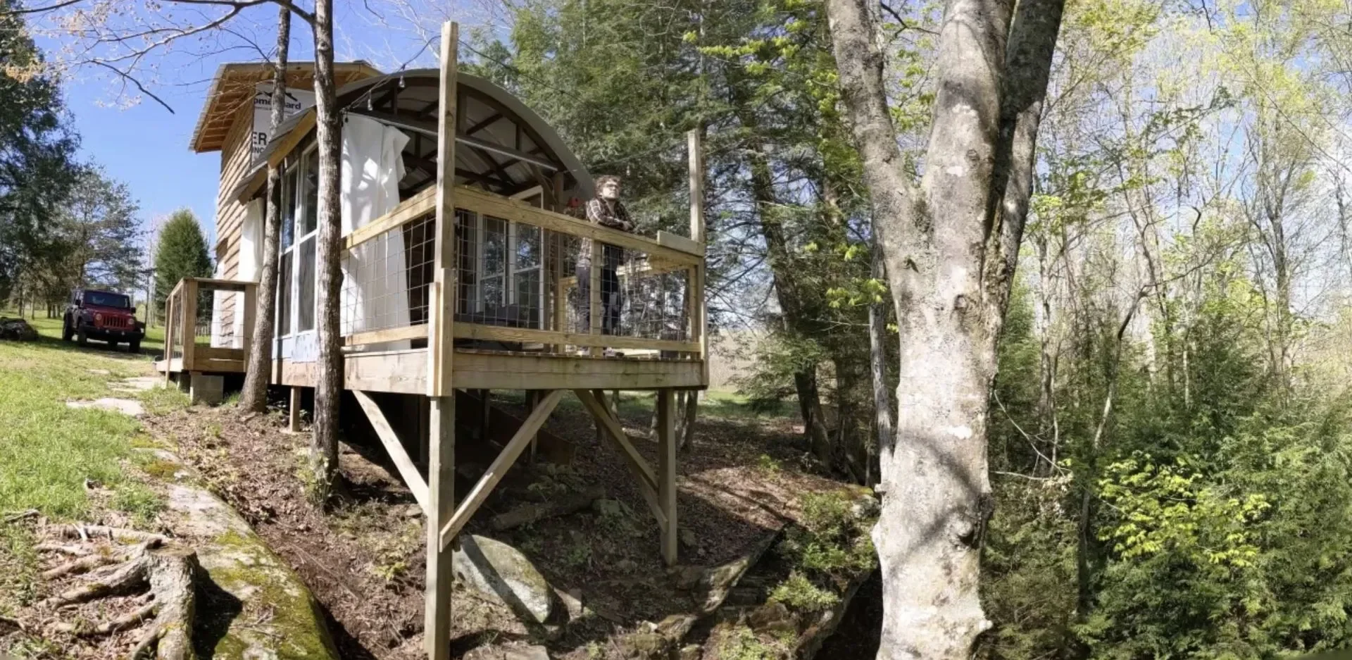A wooden treehouse with a curved roof and front porch elevated among trees, with a person standing on the porch and vehicles parked on a grassy area nearby.
