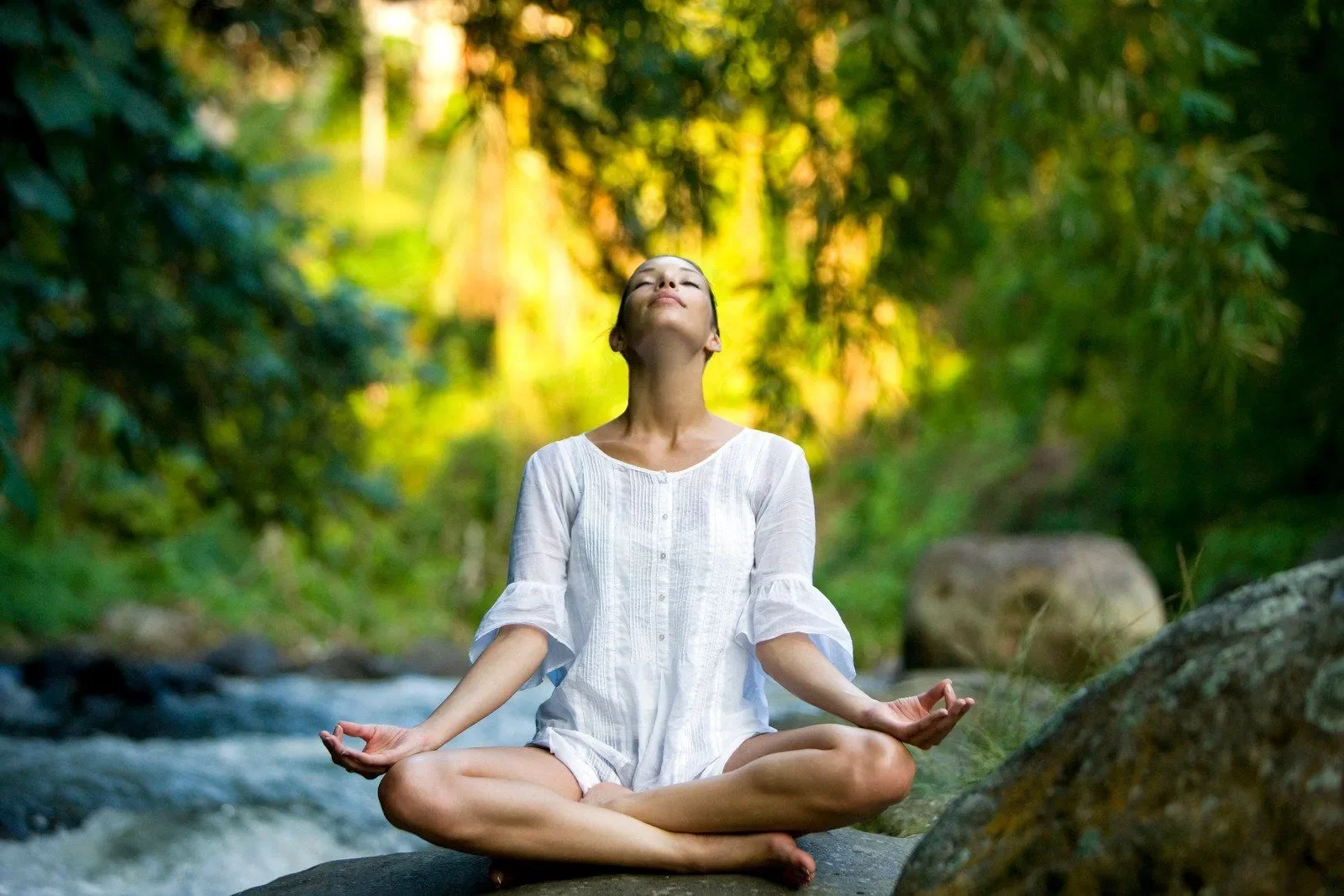 A woman practicing yoga or meditation outdoors in a serene, green forest, sitting cross-legged on a rock with her eyes closed, wearing a white dress, and appearing peaceful.