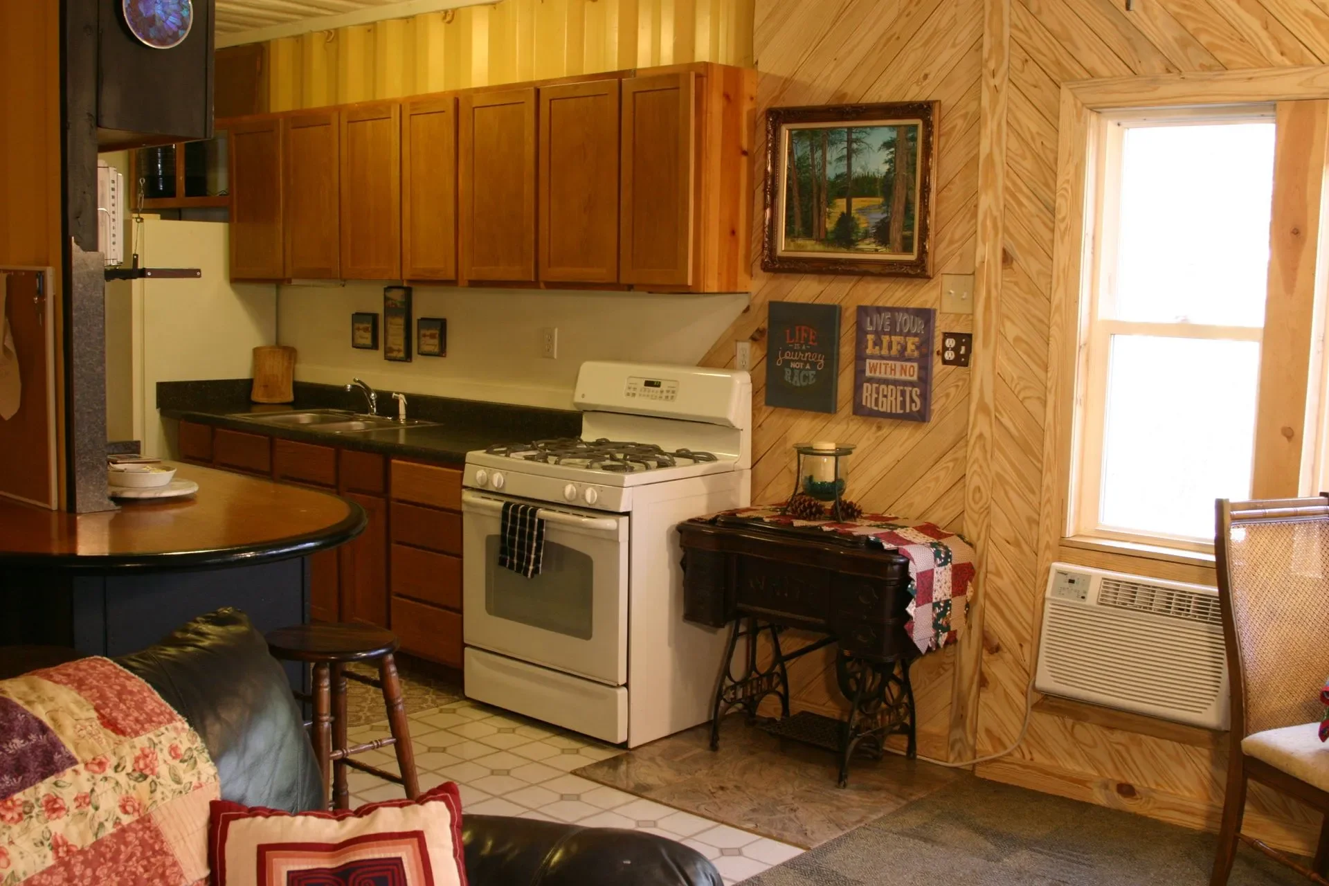 A cozy kitchen with wooden cabinets and wall paneling, a white gas stove, and a window letting in natural light. Decor includes framed art, inspiring signs, and a small vintage table.