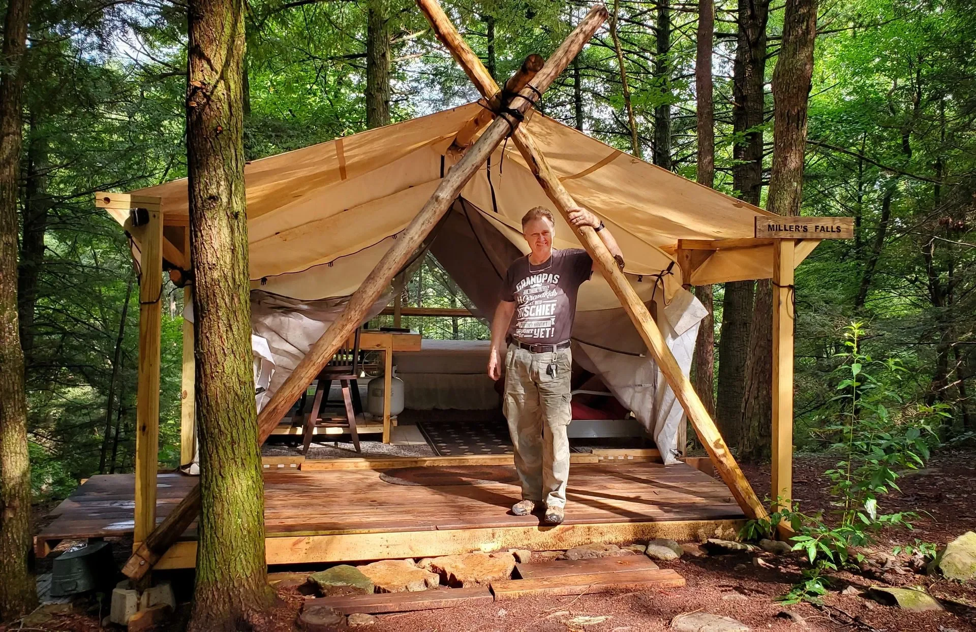 A man standing on the wooden porch of a makeshift shelter in a forest, next to a sign that reads 'Miller's Falls'. The shelter is made of wooden logs and fabric, supported by large branches, and surrounded by tall trees and green foliage.