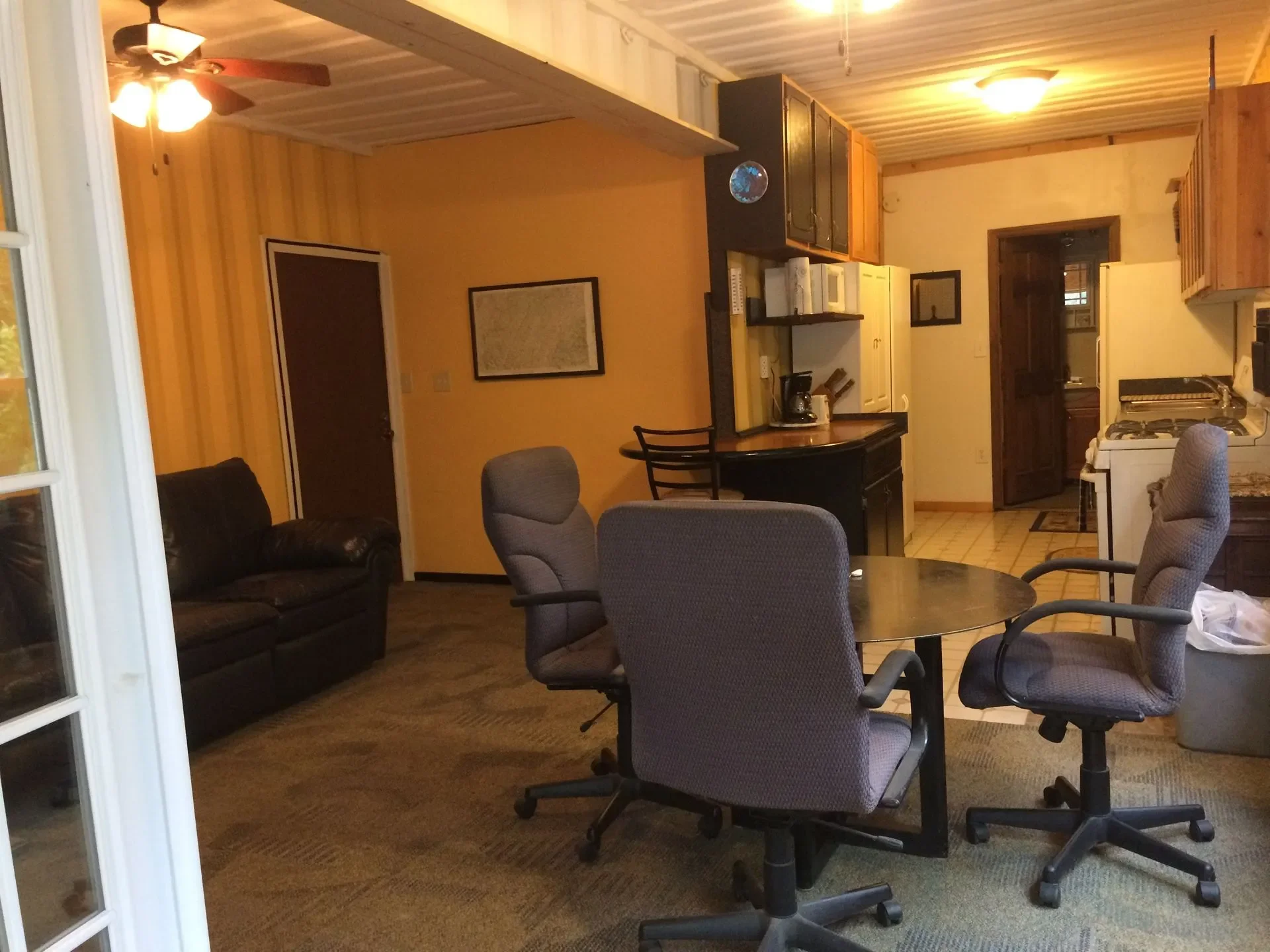 Interior of a living space with a dark leather couch, four gray office chairs around a round table, kitchen area with a coffee maker, stove, and cabinets, fan with light, and walls decorated with a framed picture.