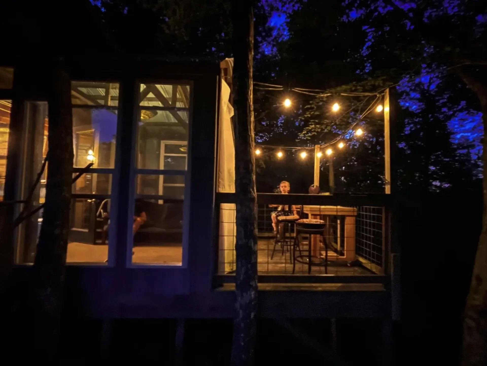 Nighttime porch scene with string lights hanging above a wooden deck. Inside, a person is sitting at a table, visible through glass windows.