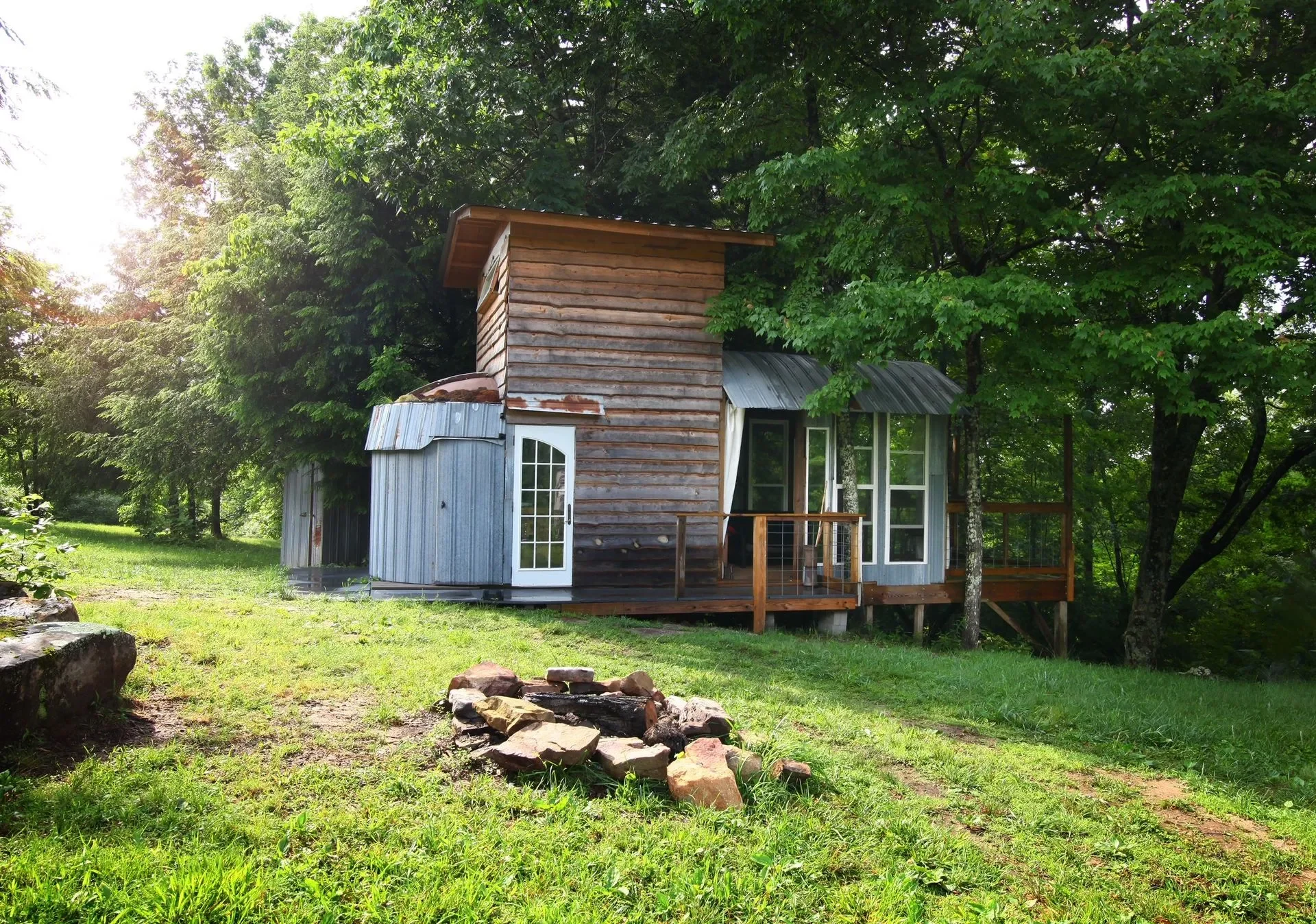 A small rustic house with a mix of weathered wood and gray metal siding, surrounded by green trees and grass, with a small stone fire pit in the foreground.