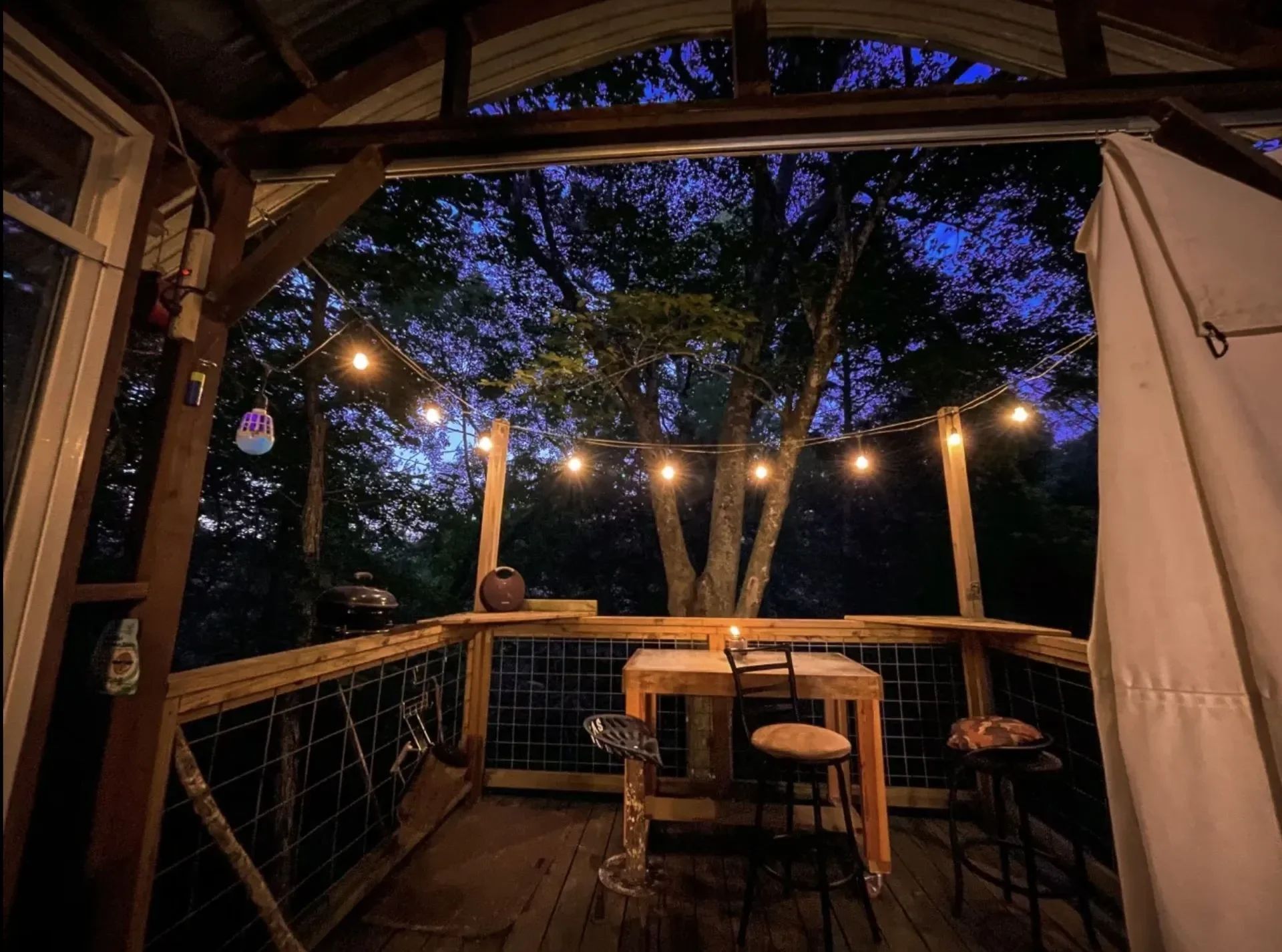 A cozy outdoor deck at night with string lights, a small wooden table, three chairs, and trees with purple twilight in the background.