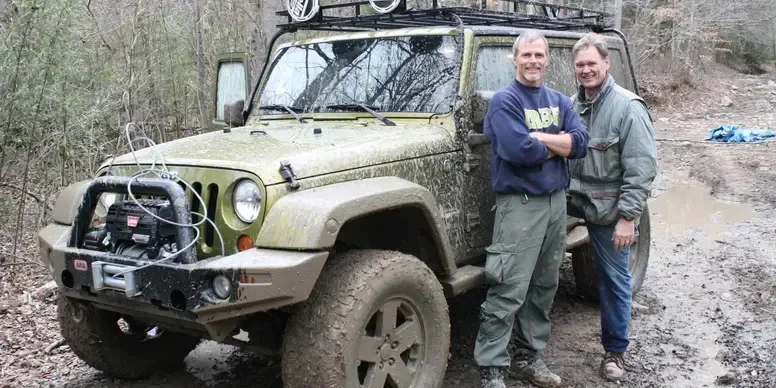 Two men standing next to a muddy off-road vehicle in the woods, smiling with arms around each other.