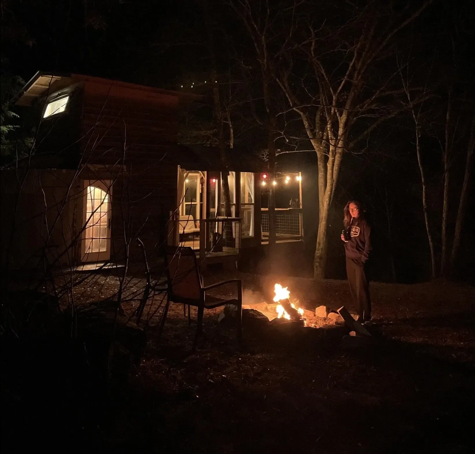 Nighttime scene showing a woman standing near a small campfire in the yard of a wooden house with lit windows, surrounded by trees.