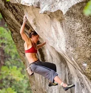 Woman rock climbing outdoors, wearing a red sports bra and gray pants, with trees in the background.