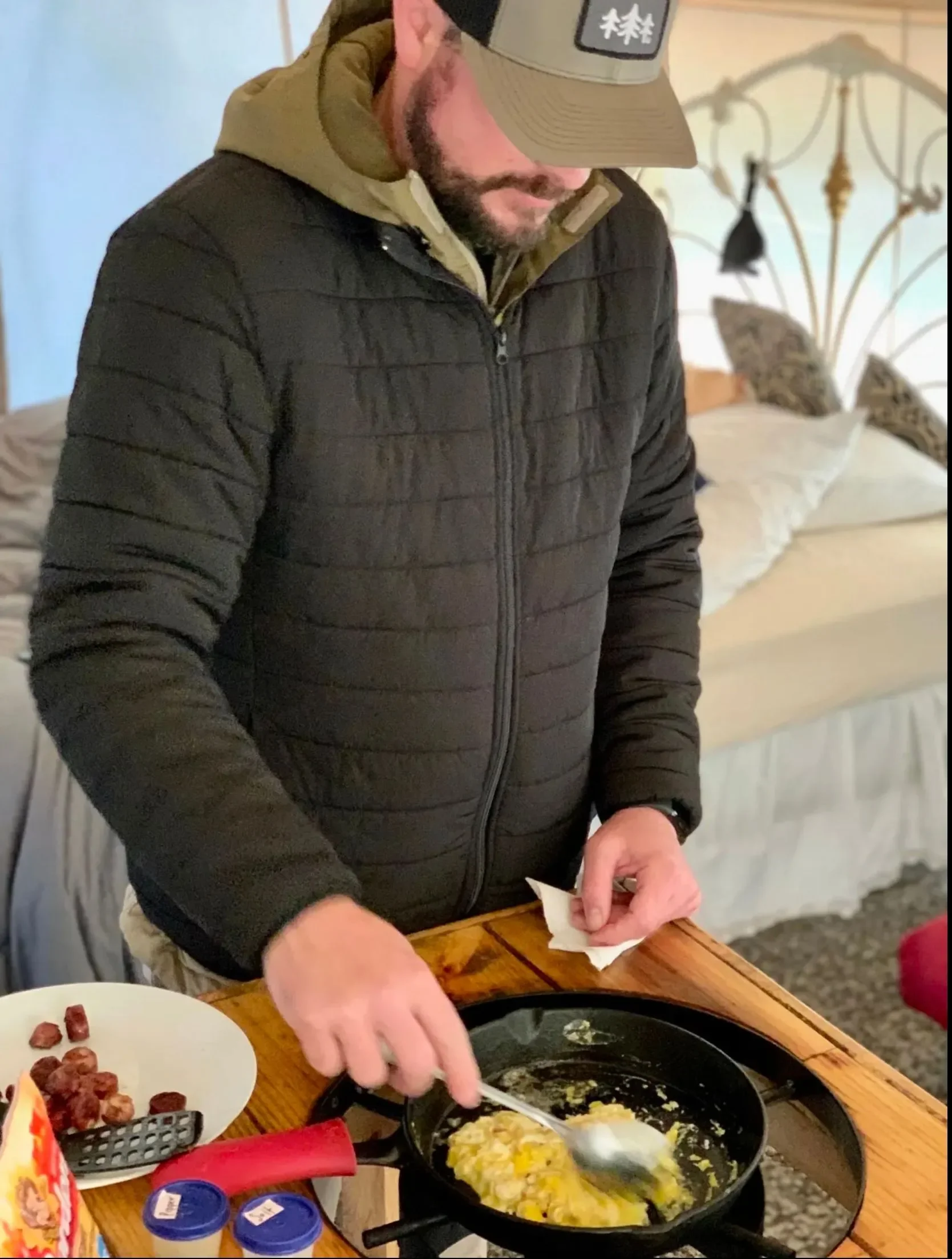 A man cooking scrambled eggs on a black skillet in a bedroom, surrounded by breakfast items.