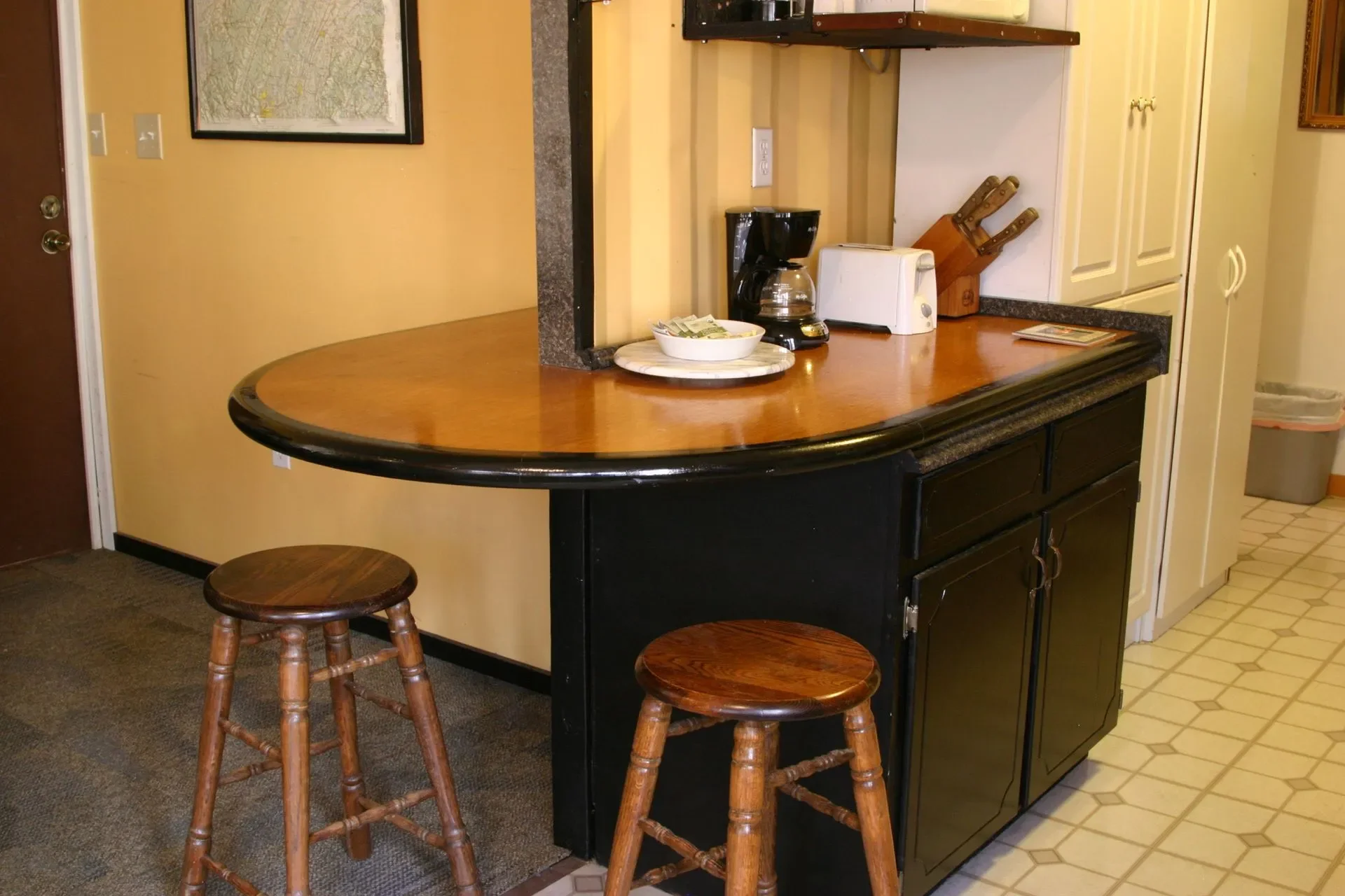 A kitchen counter with a coffee maker, toaster, and knives, with two wooden stools and a map on the wall.