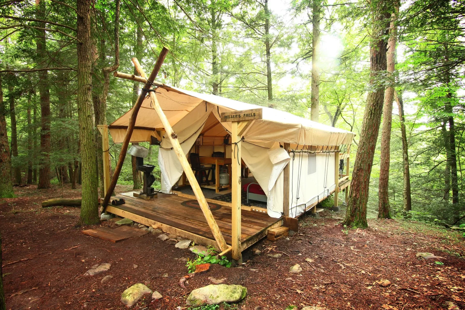A rustic tent set up on a wooden platform in a forest with trees, greenery, and sunlight filtering through the leaves.