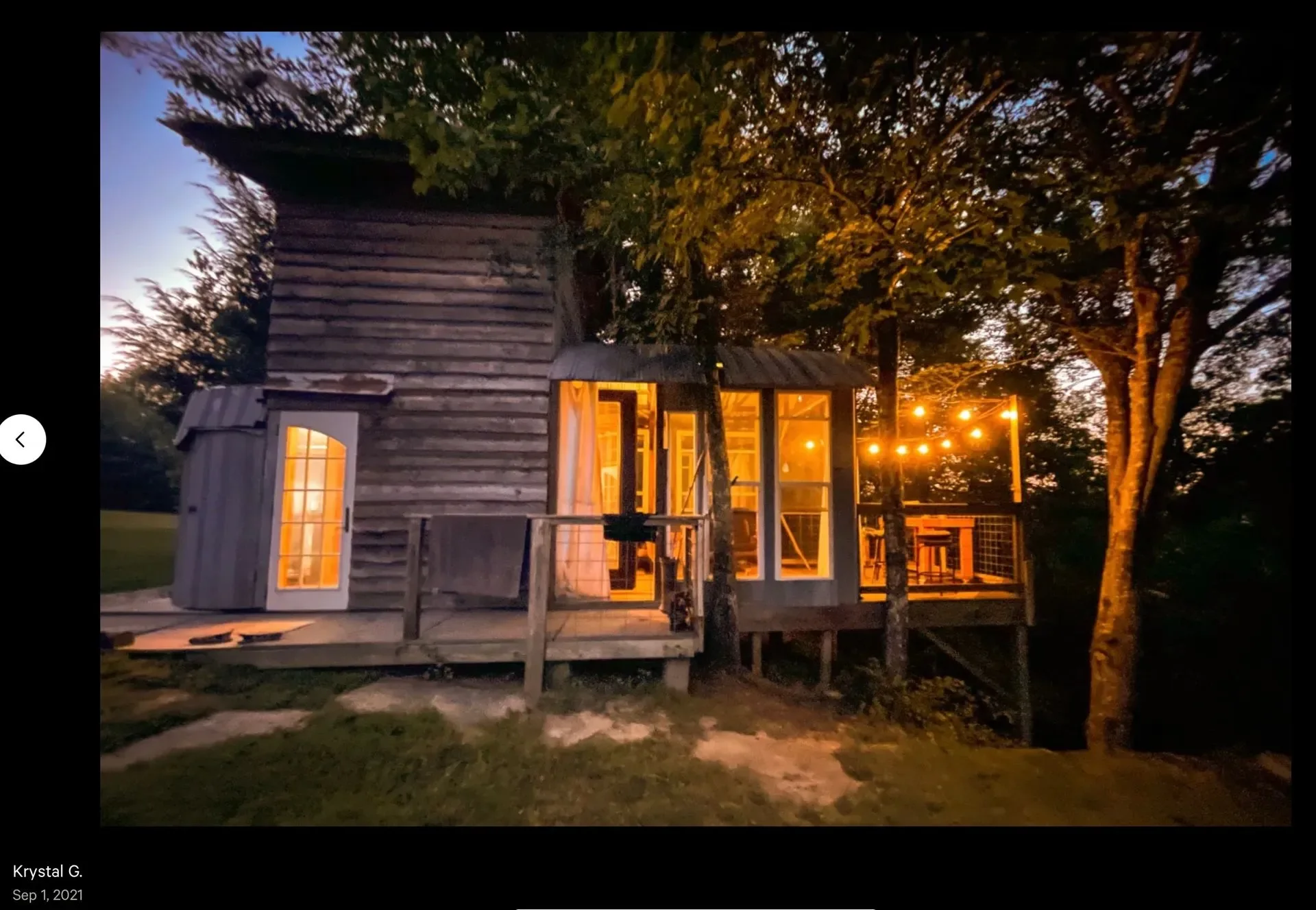 A small wooden house with large windows and a screened porch illuminated by warm lighting, surrounded by trees at dusk.