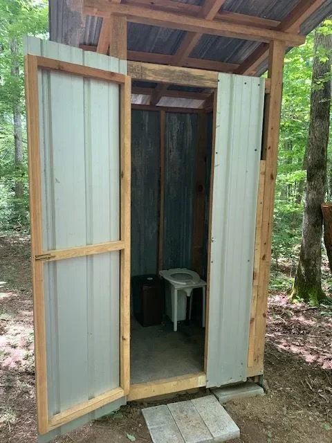 A small outdoor toilet structure with a door open, showing a squat toilet inside, situated in a wooded area.
